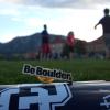 A Be Boulder pin sits atop poster paper on a field on campus with people in the background.