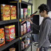 Man wearing blindfold holds up a camera on a walking stick to scan a shelf containing several different brands of cereal.