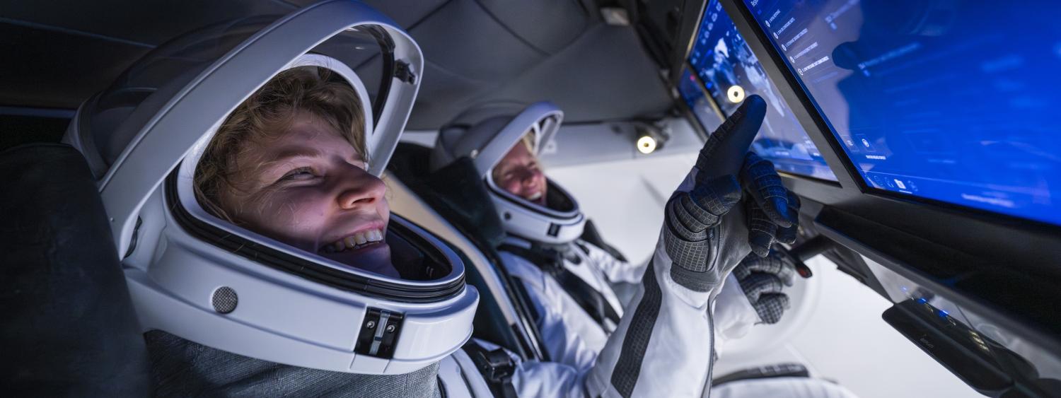 Two astronauts wearing open helmets sit inside a cockpit