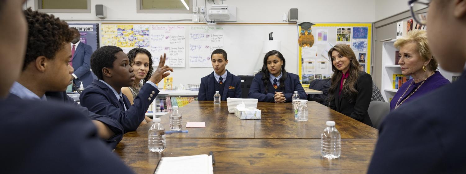 School children in blue blazers sit around a large table speaking to woman in a blue pantsuit