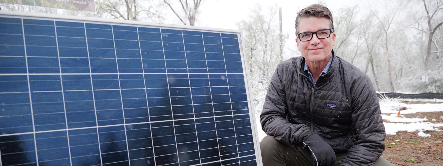 Man kneels on the ground next to a large solar panel with snow in the background