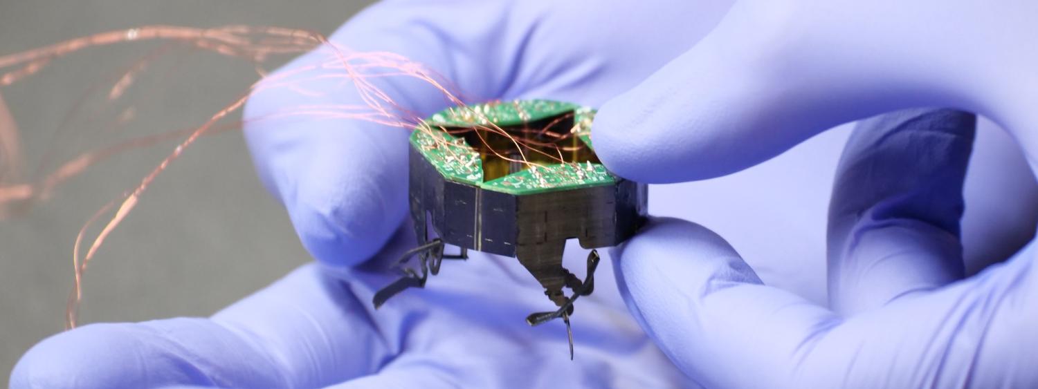 Hands wearing purple gloves hold a tiny robot with wires running from it