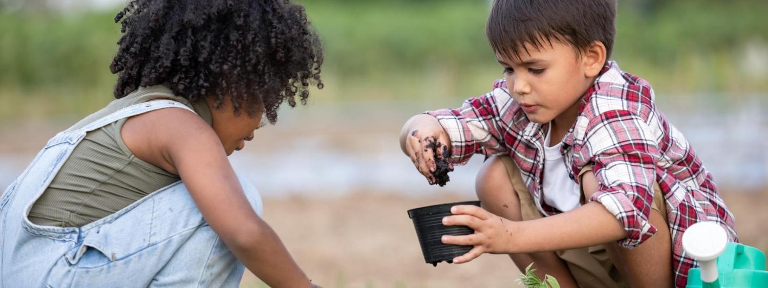 Two children kneel in the grass, scooping piles of dirt into plastic pots