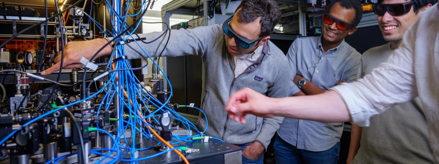 Four men gather around a lab table that is covered in machinery, lenses and wires.
