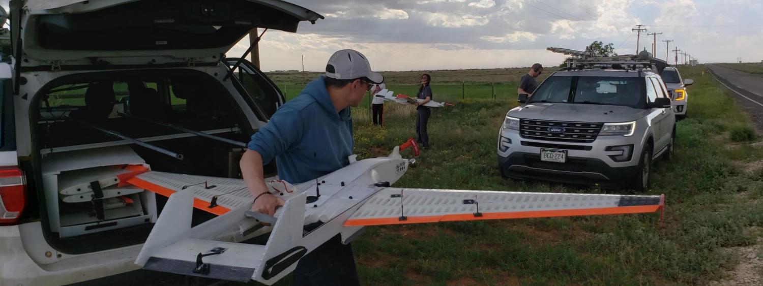 Man lifts drone with fixed wings out of the trunk of an SUV while other scientists work in background, and storm clouds brew above