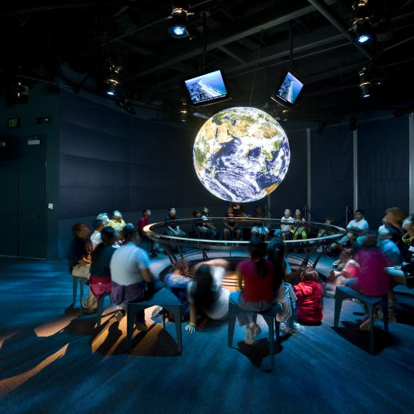 Children sit in a circular gallery around a glowing, suspended model of Earth, participating in an interactive science presentation inside a darkened museum space.