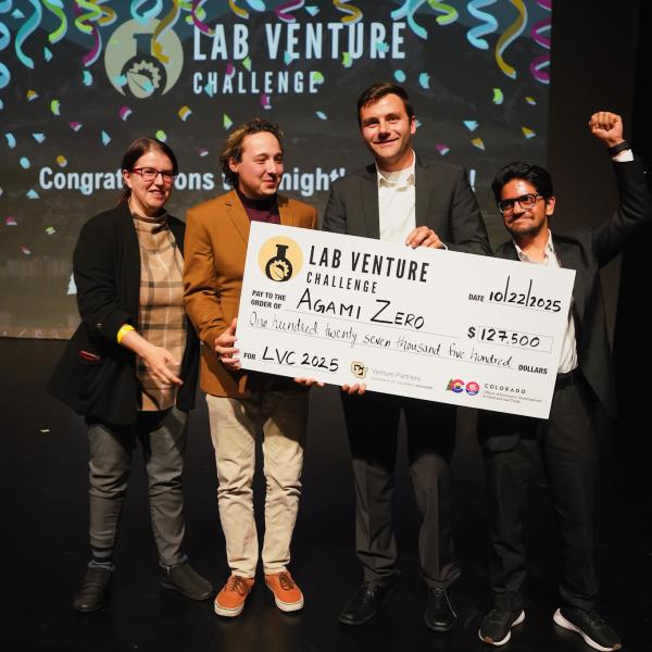 Four people stand on stage celebrating as they hold an oversized check for $127,500 made out to Agami Zero, winners of the 2025 Lab Venture Challenge, with confetti graphics and the event logo projected behind them.
