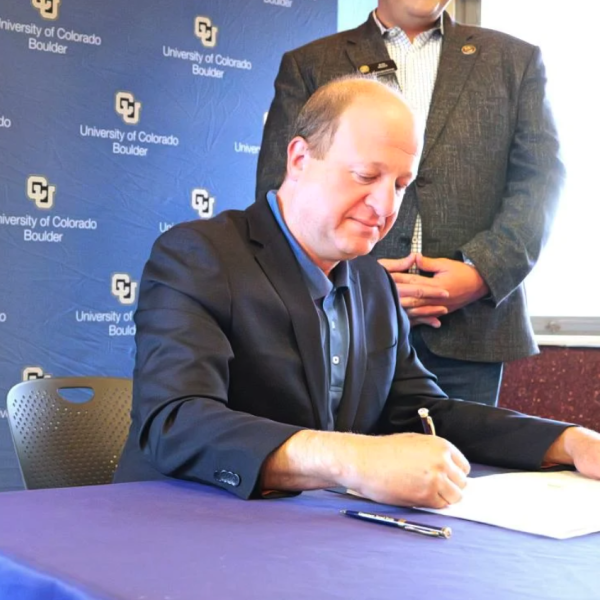 A balding man in a suit sits at a desk in front of the CU Boulder logo and the official seal for the State of Colorado signs a document