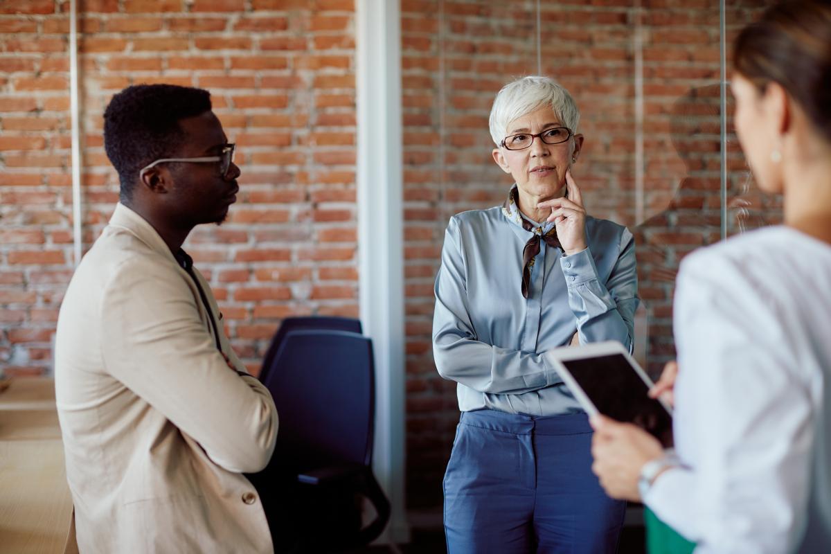 A black man in a tan suit leans on a desk listening to an older white woman in a blue blouse talk to a younger white women in a white blouse holding a tablet.