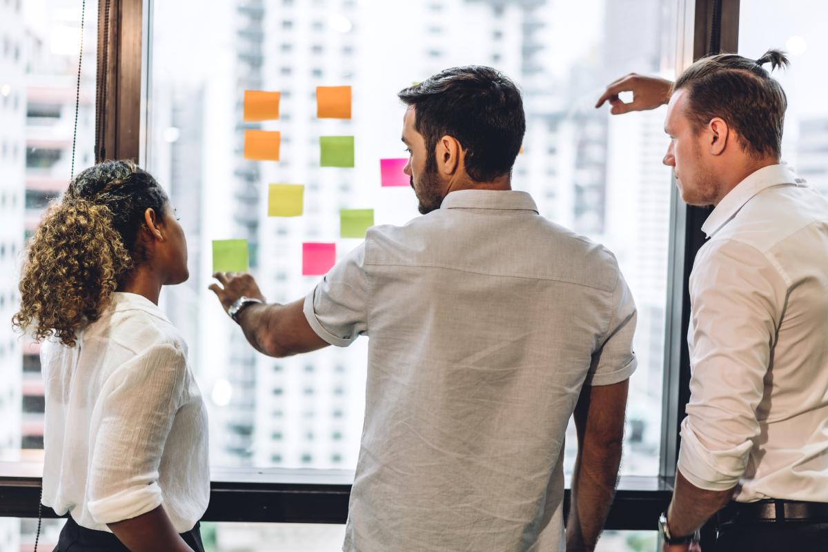 Two white men and a woman of color work on a strategy with sticky notes on a winodw
