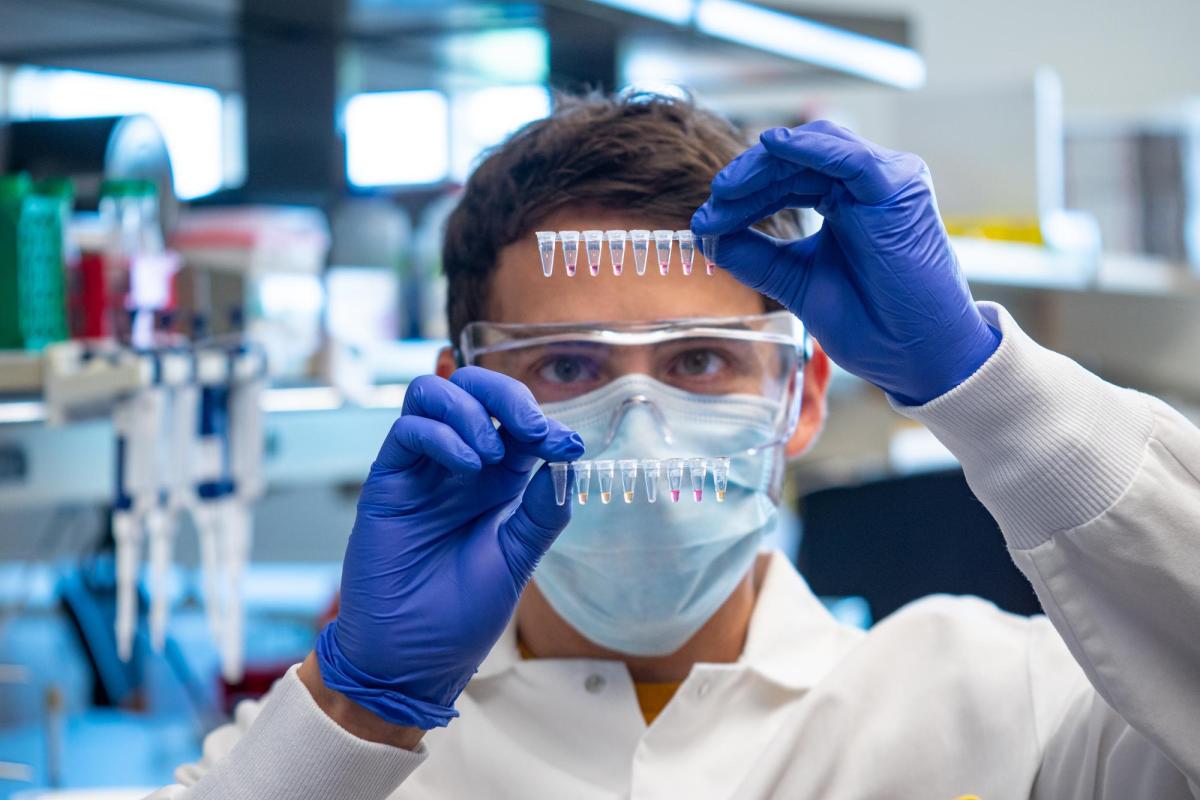 A white male researcher with short brown hair wearing a face mask and protective glasses holds up tiny vials.