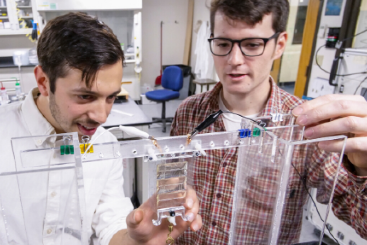 Two white men in button-up shirts work on a structural device in a lab