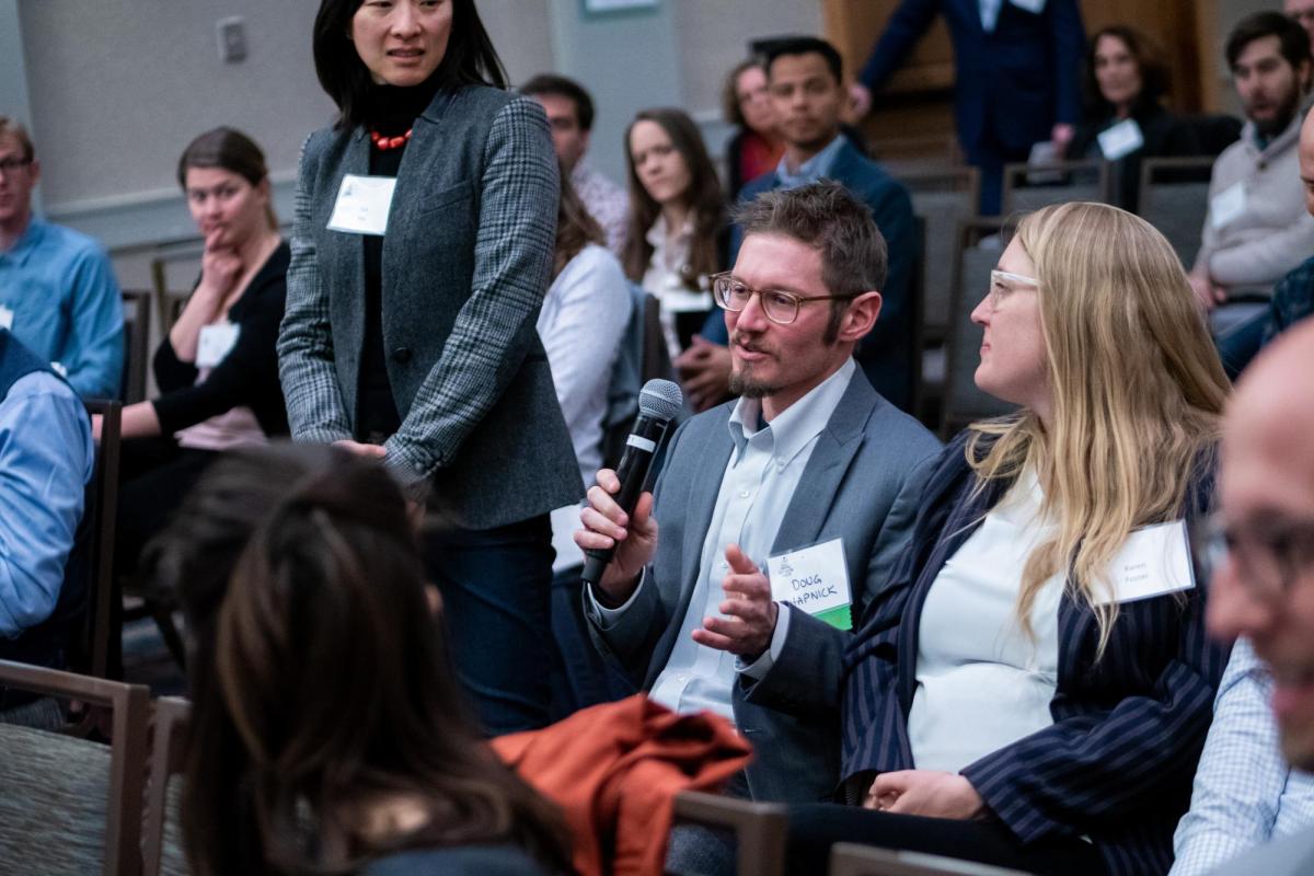 A diverse group of business people in a the audience of a presentation; a man wearing glasses and a grey suit speaks into the microphone.