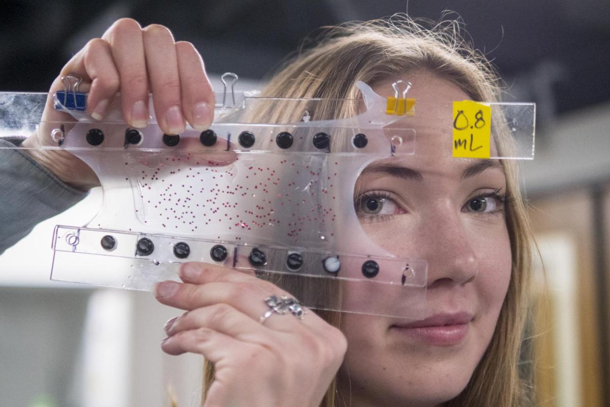 A blonde, white female researcher holds up two pieces of plastic between which is a stretched transparent material she looks through with one eye.