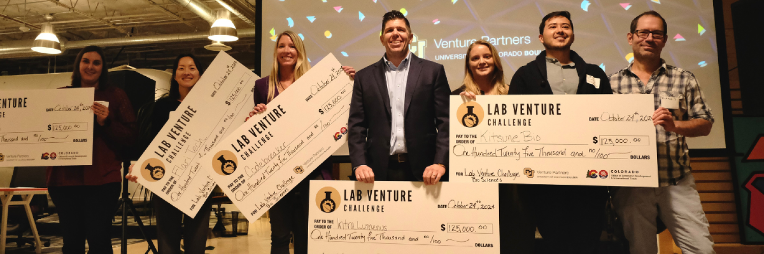 A diverse group of people stands on a presentation stage holding giant prize checks.