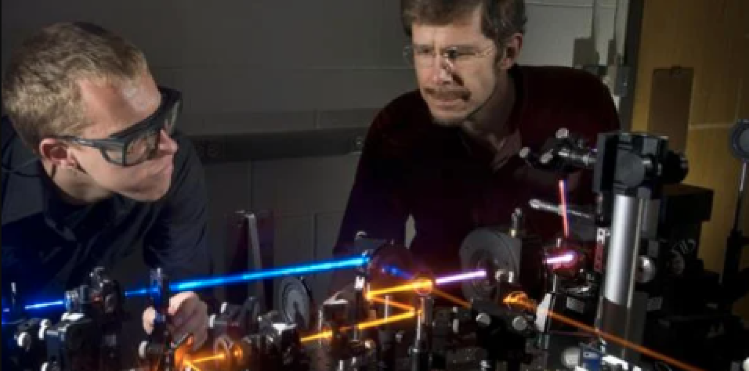 The image shows two masculine researchers in a well-equipped electrical engineering lab. They are gathered around a workbench, intently focused on their projects. The lab is filled with various electronic components, tools, and equipment.
