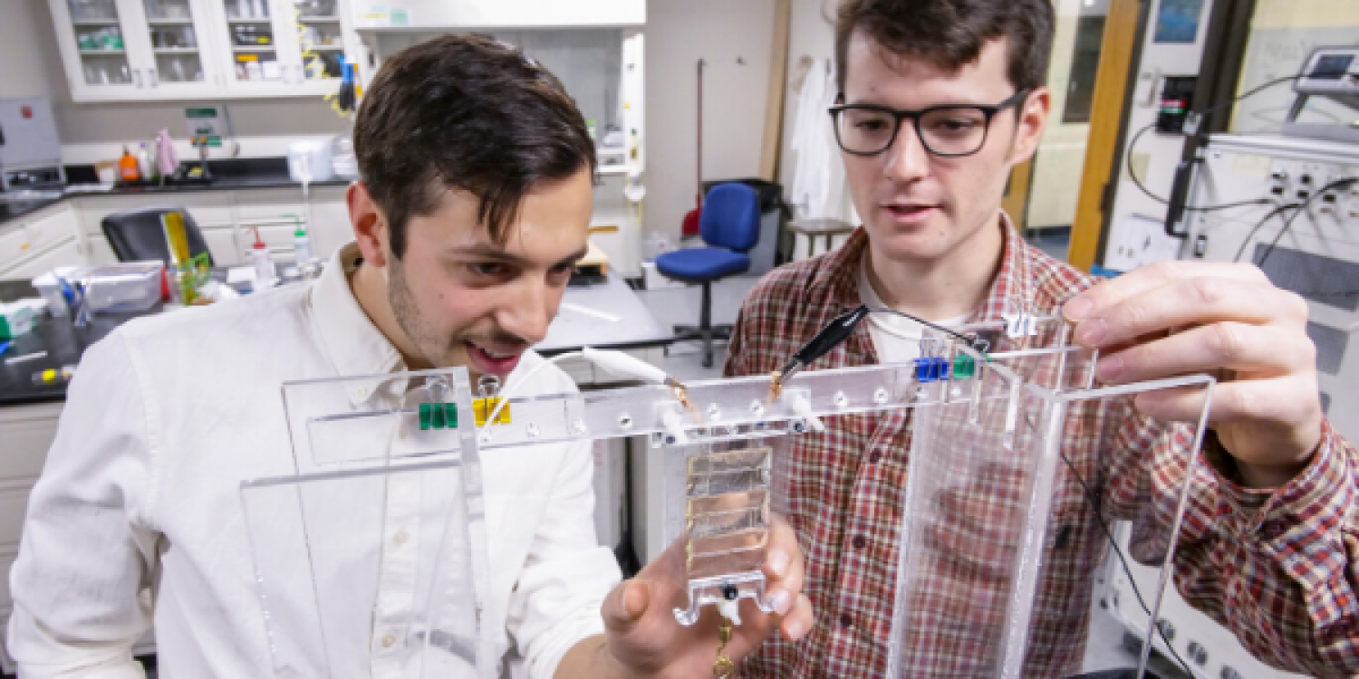 Two white men in button-up shirts work on a structural device in a lab