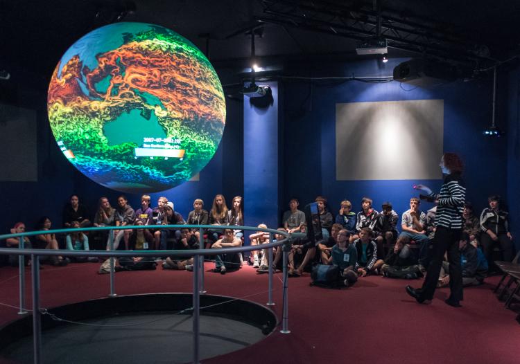 hildren sit in a circular gallery around a glowing, suspended model of Earth, participating in an interactive science presentation inside a darkened museum space.