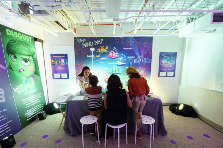 Children and an adult gather around an interactive exhibit titled &lsquo;Riley&rsquo;s Mind Map,&rsquo; exploring emotions through hands-on activities in a brightly lit gallery inspired by the film Inside Out.