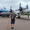 A man wearing a CU Boulder athletics jersey stands in front of a storm-chasing airplane on an airport tarmac.