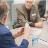 Men sit at a table discussing the device one man holds in his hands.