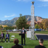 An outdoor event with attendees wearing business suits sitting on a green field next to a tall white tower.