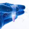 A hand wearing blue medical gloves holds up a small vial with an illustration of DNA superimposed on the image.