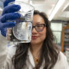 A woman with long dark hair and black glasses holds up a beaker to the camera with a jellyfish floating inside of it.