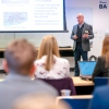 A man stands and gives a presentation to a group of people in a classroom
