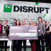 Alternate text: “A group of startup founders and event organizers stand on a stage beneath a large ‘DISRUPT’ sign, holding a giant symbolic check for $100,000 labeled ‘Startup Battlefield 200’ and celebrating with confetti falling around them.”