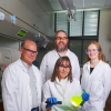 Four researchers wearing lab coats and safety glasses pose in a laboratory; the seated scientist holds a glowing yellow-green flask illuminated under a blacklight, while the others stand smiling behind her.