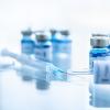 Close-up of medical syringes and vials filled with blue liquid labeled ‘vaccine’ arranged on a reflective surface in a bright laboratory setting.