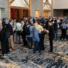 A large group of people networking and conversing in a spacious hotel conference foyer outside the Adams Ballroom during an event. Attendees wearing business casual attire stand in small clusters around tall tables, holding coffee cups and badges, while others mingle in the background under warm lighting.