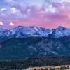 Panoramic view of snow-capped Rocky Mountains at sunrise, with green pine forests and open valleys below under a pink and purple cloud-filled sky.