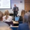 A speaker stands at the front of a classroom-style lecture hall, presenting slides on quantum engineering to an audience seated at tables with laptops and notebooks.