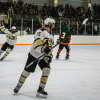 The image shows a fast-paced moment during a CU Buffs hockey game, with a player in a black and gold jersey gliding swiftly across the ice. The player has the puck on their stick, closely pursued by a member of the opposing team in a white and blue jersey. Both players are focused on the action, surrounded by the bright rink lighting and the distant blur of the crowd in the background. The CU Buffs logo is visible on the player's jersey, symbolizing the team's energy and intensity during the game.