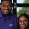 The image shows two individuals, a Black man and a South Asian woman, both wearing purple shirts with the "MesaQuantum" logo printed on the left side. They are smiling and facing the camera. The man, on the left, has short hair and a beard, while the woman, on the right, has long black hair and is wearing pearl earrings. The background features large windows with greenery outside, creating a bright, welcoming, and professional setting.