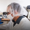 A man wearing protective lab clothes looks through a microscope