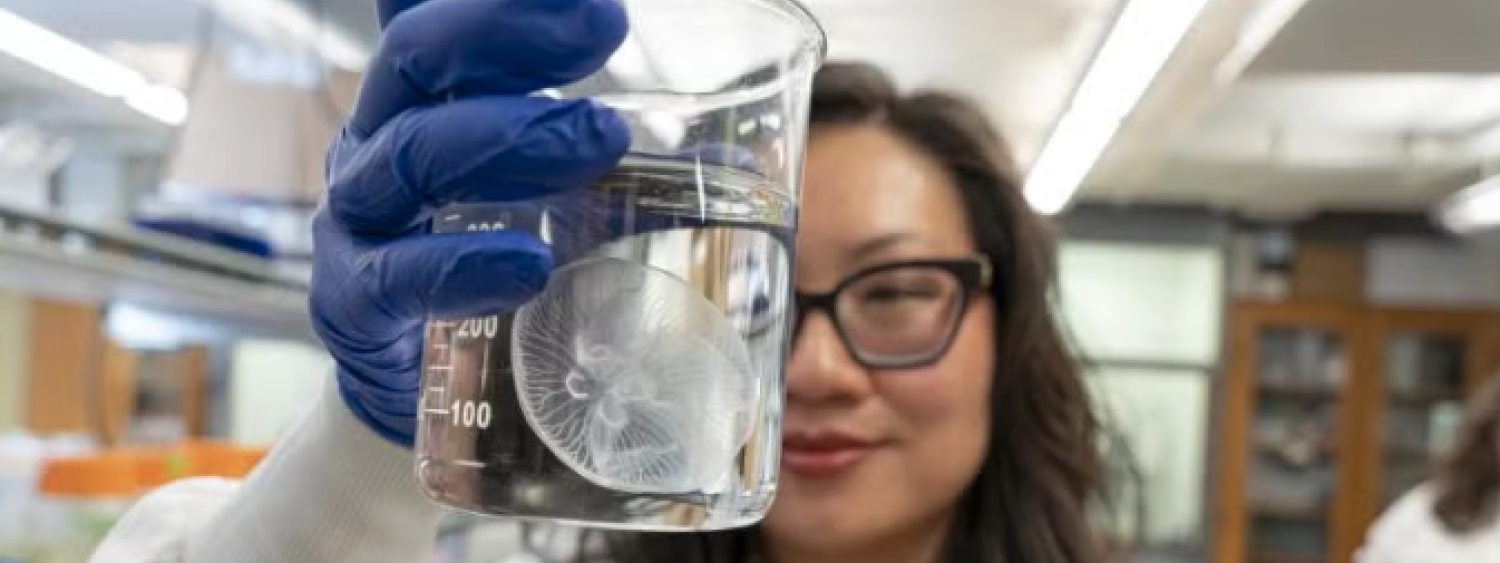 A woman with long dark hair and black glasses holds up a beaker to the camera with a jellyfish floating inside of it.