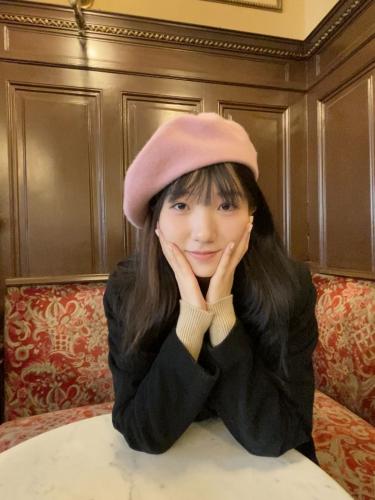 A person with bangs and long hair wearing a pink hat smiles with their hands on their chin