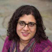 Professor Samira Mehta smiles up at a camera while sitting. Mehta wears a magenta sweater, black shirt, scarf, and glasses, and has shoulder length brown curly hair.