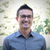 Prof Emmanuel David Smiles wide in front of a Green grassy background. David is wearing glasses and a blue button up and has short dark brown hair and