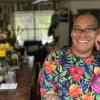 Dr. Moore is pictured in a bright floral and foliage printed dress with short sleeves and is wearing black rimmed glasses sitting with her hand up against her cheek in front of a blurred background of yellow flowers on a counter with a large picture window in the background.